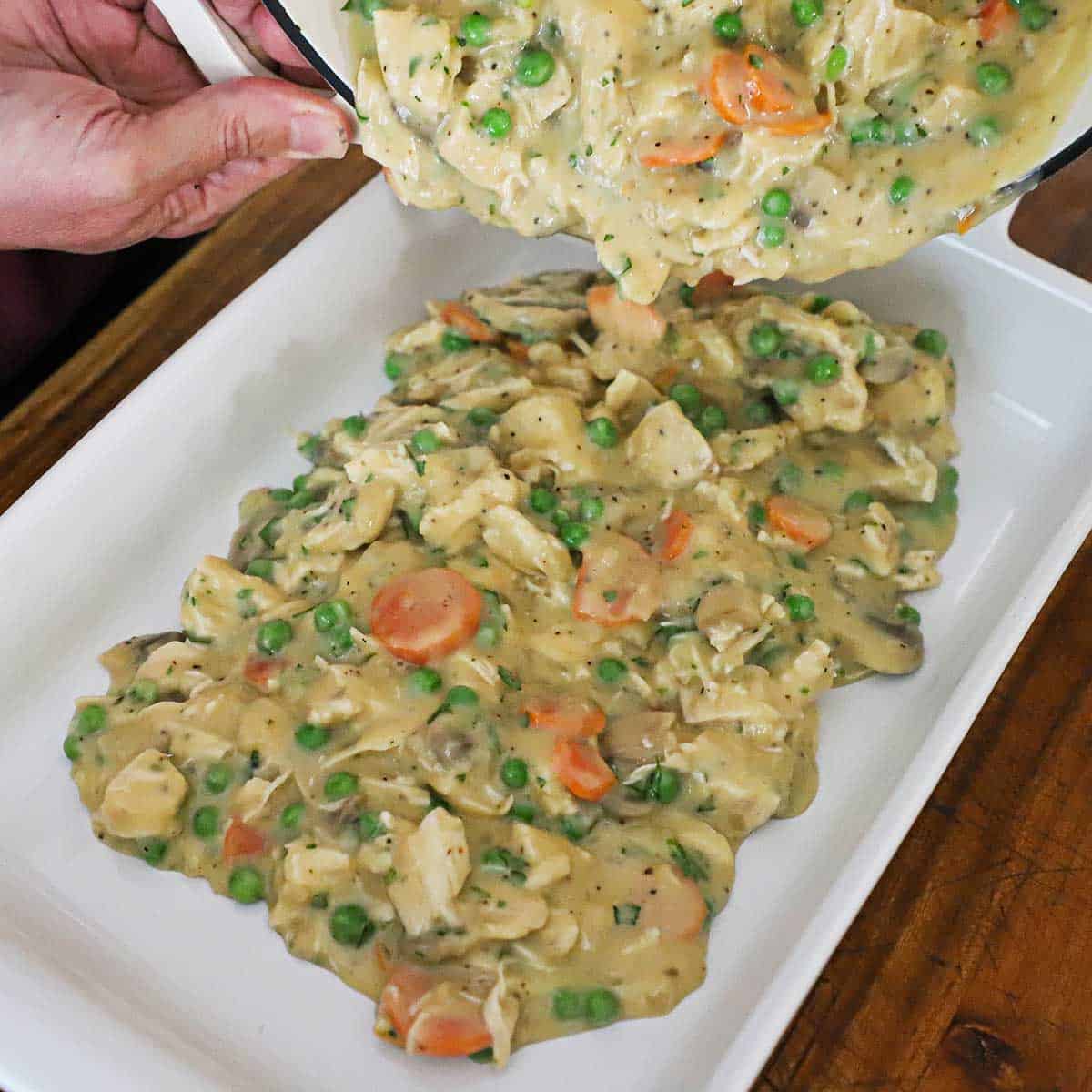 A person pouring chicken pot pie filling into a 9 by 13 white baking dish.