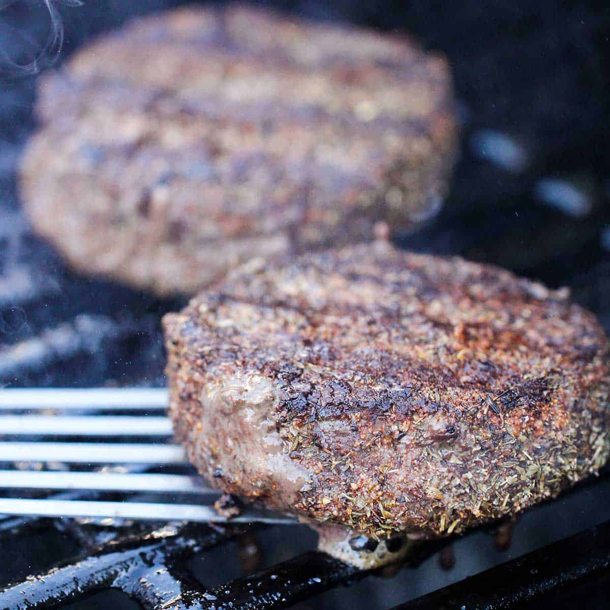 Two blackened hamburger patties that are being cooked on a grass grill.