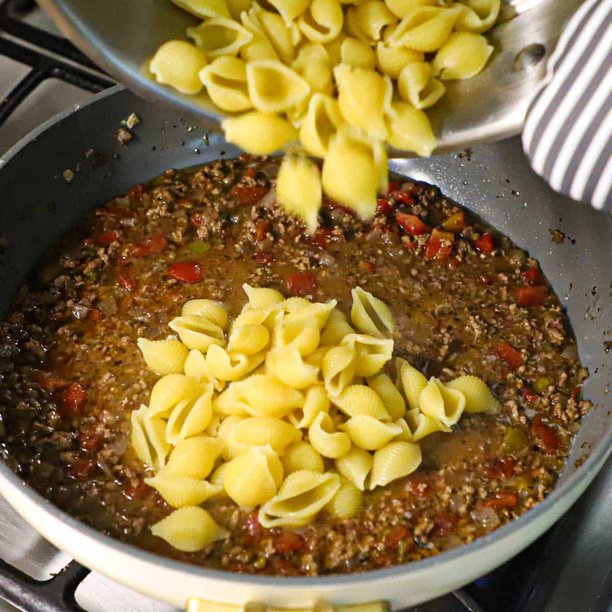 A person transferring cooked pasta shells from a pot into a skillet that is filled with a simmering meat and tomato sauce.