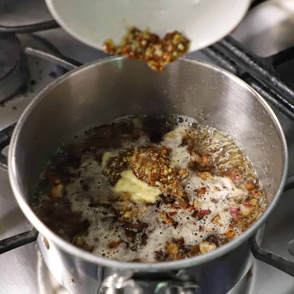 A person transferring stone-ground mustard from a small bowl into a silver saucepan that is filled with simmering chicken broth, vinegar, and seasonings.