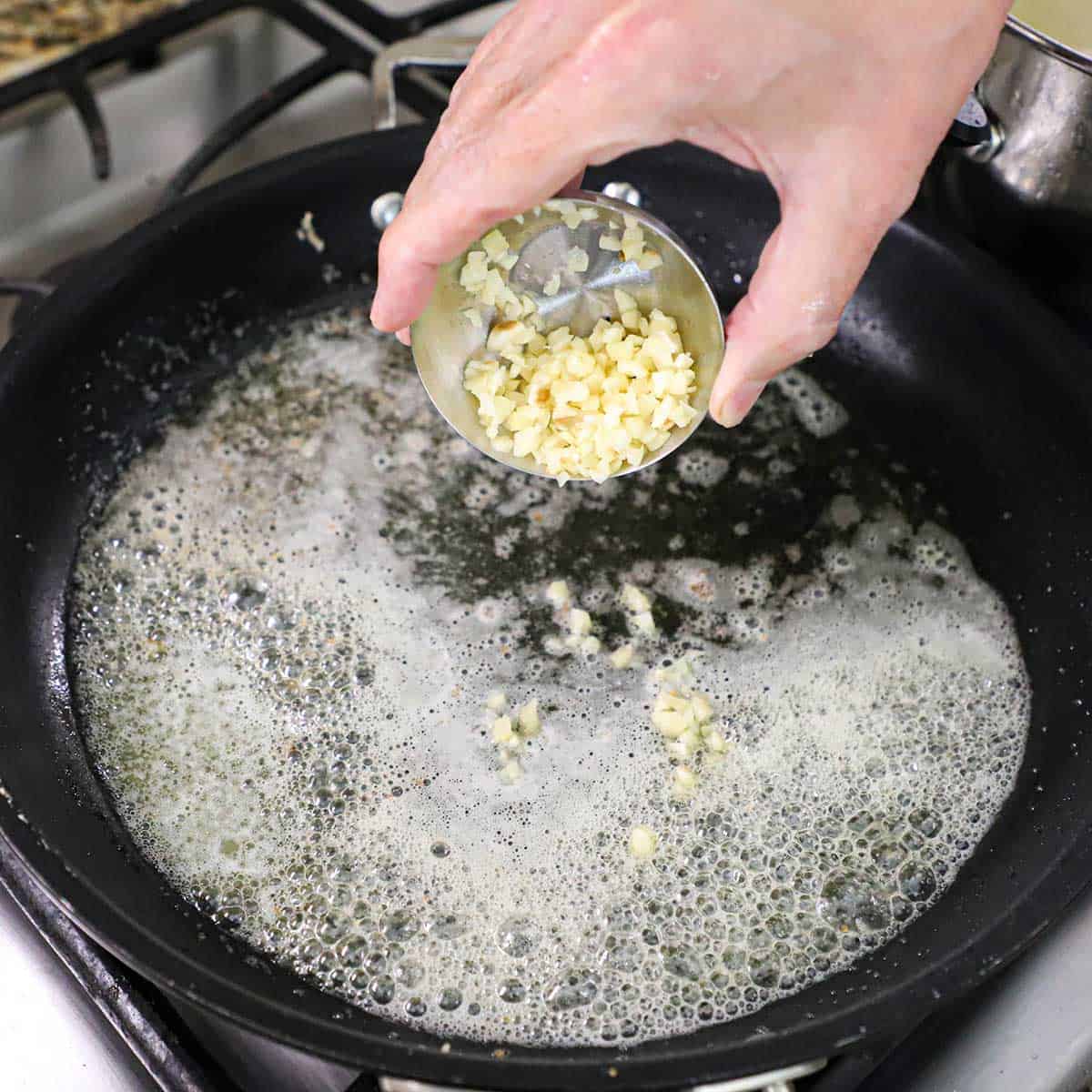 A person adding diced garlic from a small silver bowl into a large black skillet filled with simmering butter and olive oil on a gas stove.