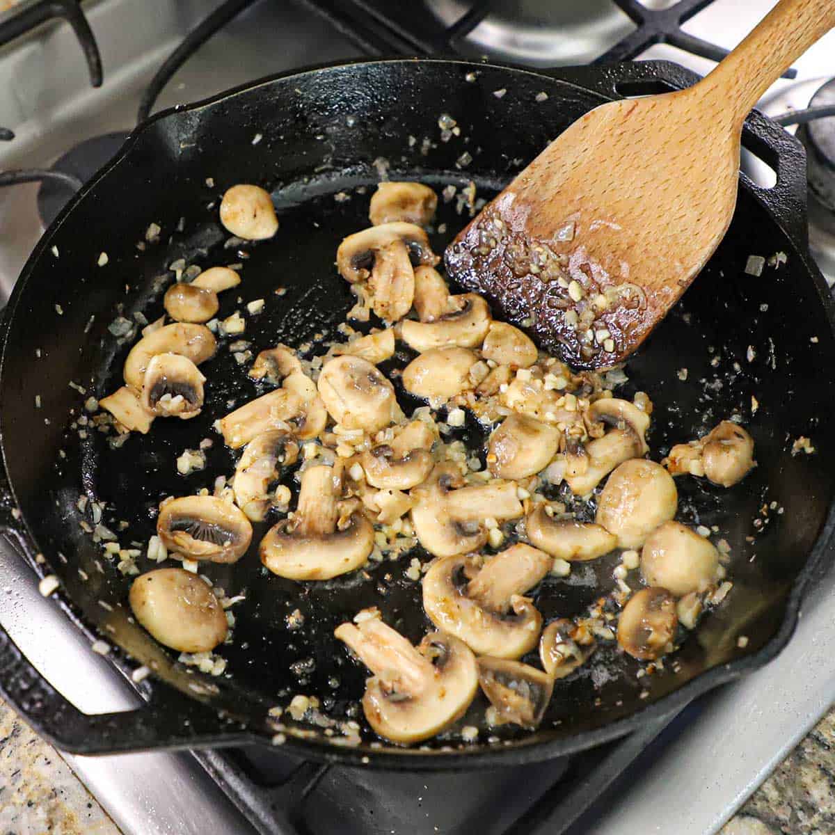 A person using a wooden spatula to stir sliced mushrooms and chopped garlic that are being sautéed in butter in a large black cast-iron skillet.