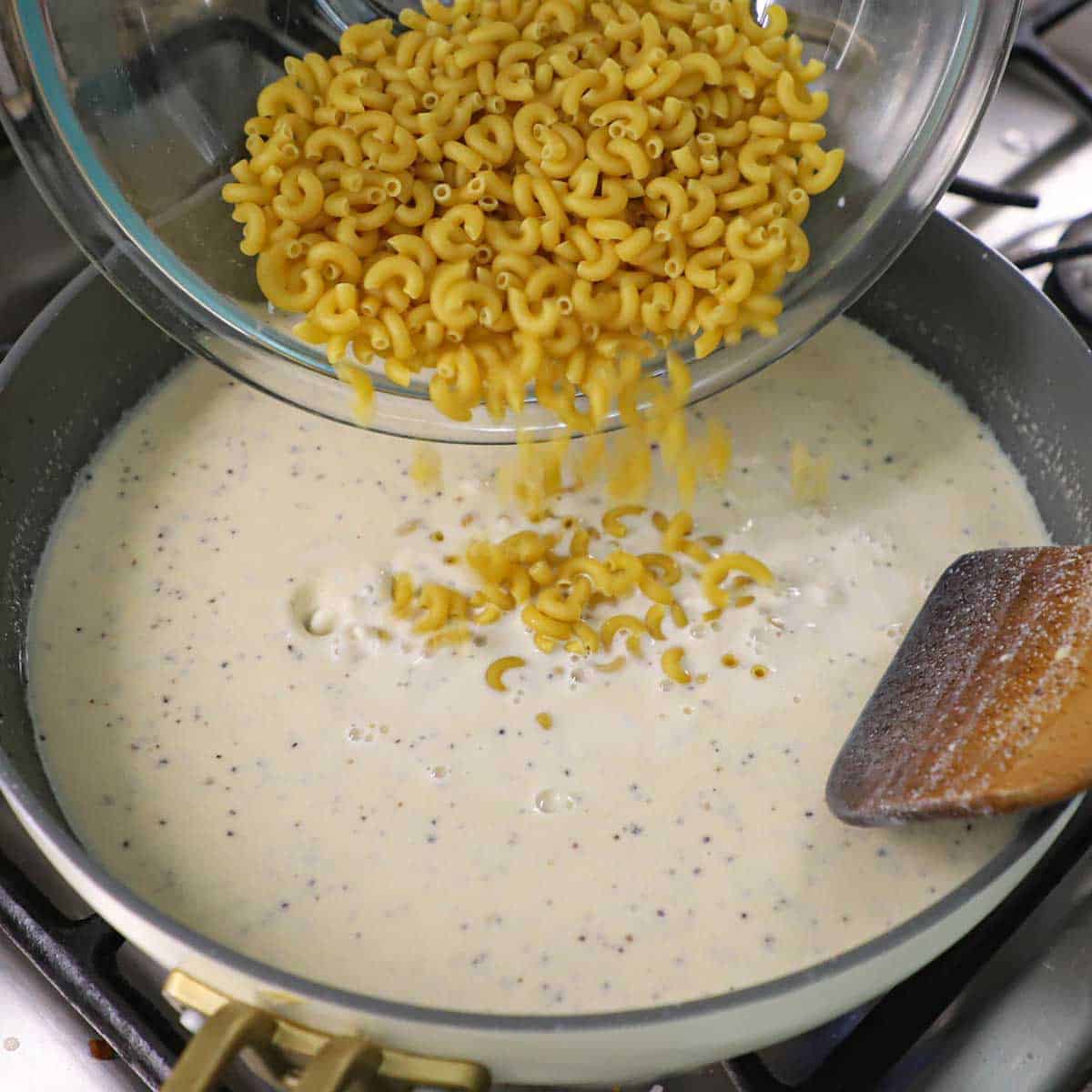 A person transferring uncooked small elbow pasta from a large glass bowl into a skillet filled with a creamy bechamel sauce.