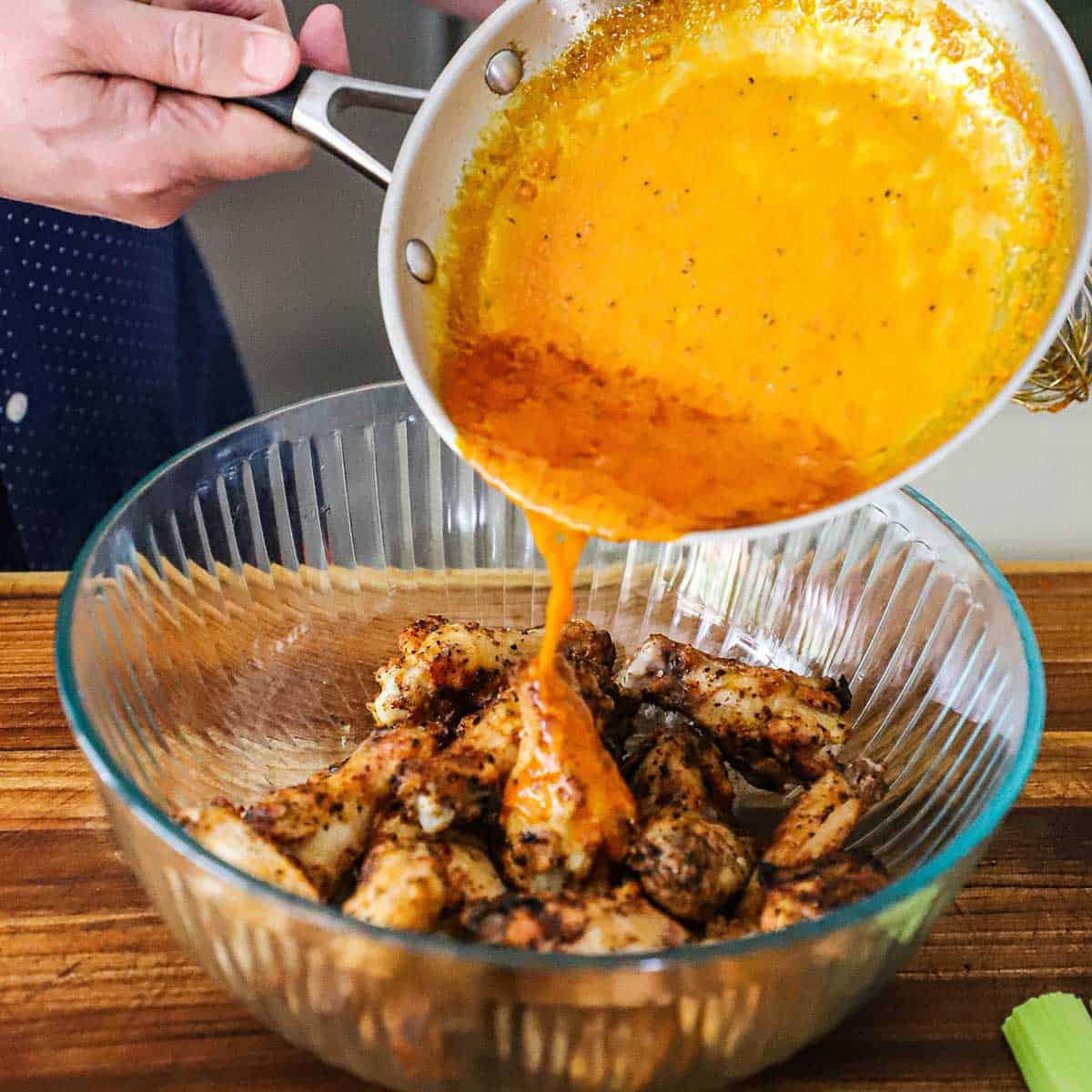 A person pouring buffalo wings sauce from a skillet into a glass bowl filled with grilled chicken wings.