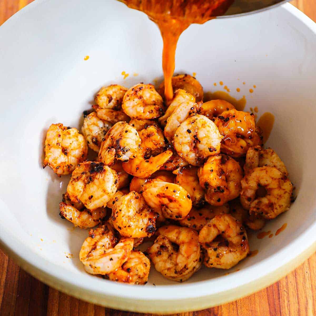 Warm buffalo sauce being poured from a saucepan into a ceramic bowl that is filled with grilled shrimp.