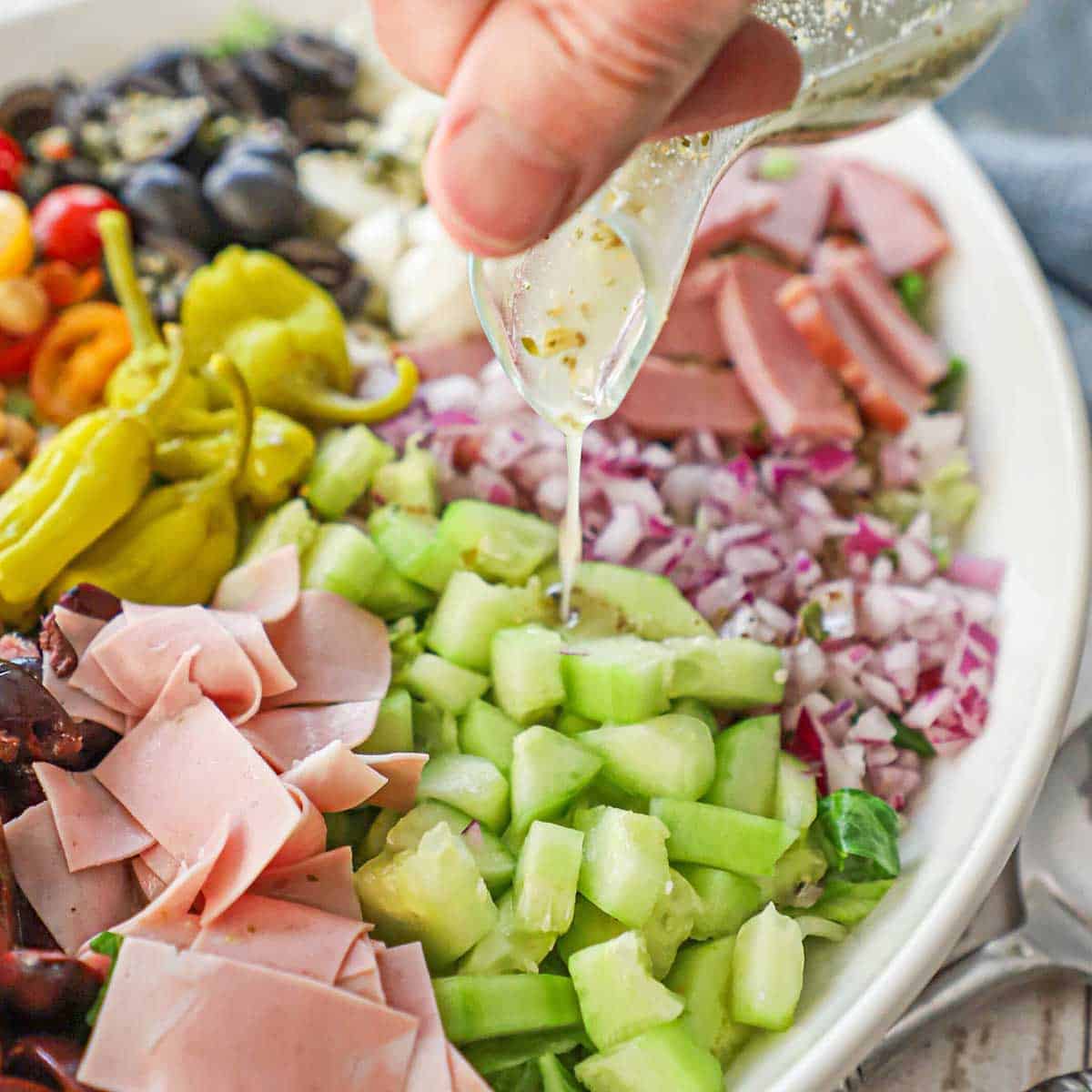 A person pouring Italian dressing from a small glass pitcher over a platter filled with the components for Italian chopped salad.