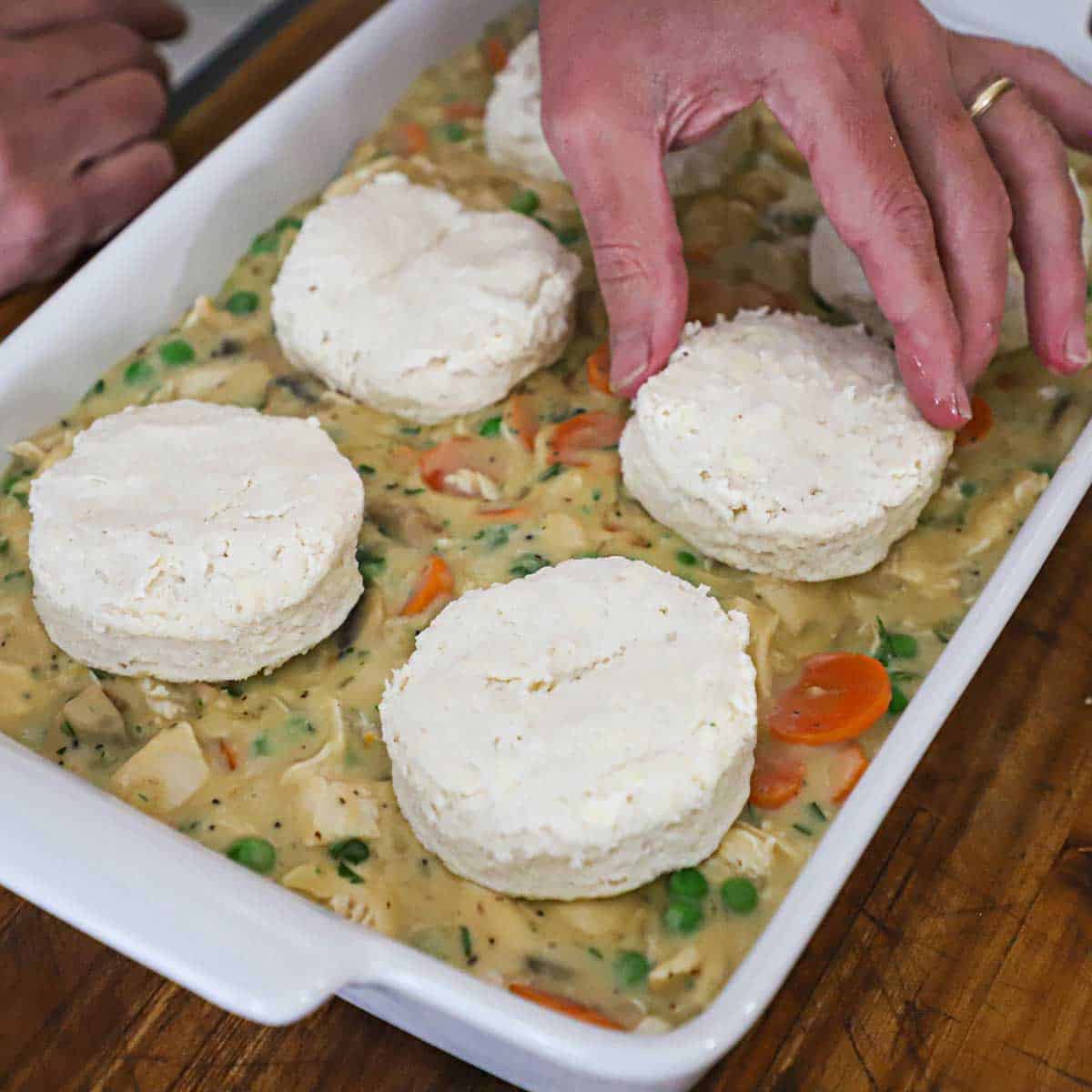 A person placing uncooked buttermilk biscuits over the top of chicken pot pie filling in a 9 by 13 white baking dish.