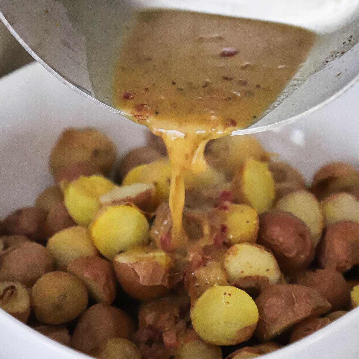 A warm bacon vinaigrette being poured over a piled of partially peeled and fully cooked baby potatoes in a white serving bowl.