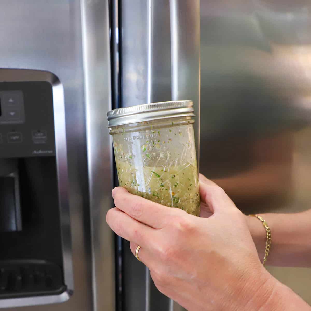 A person holding a glass jar with lid that is filled with homemade Italian dressing in one hand and is getting ready to open the door of the refrigerator with his other hand.