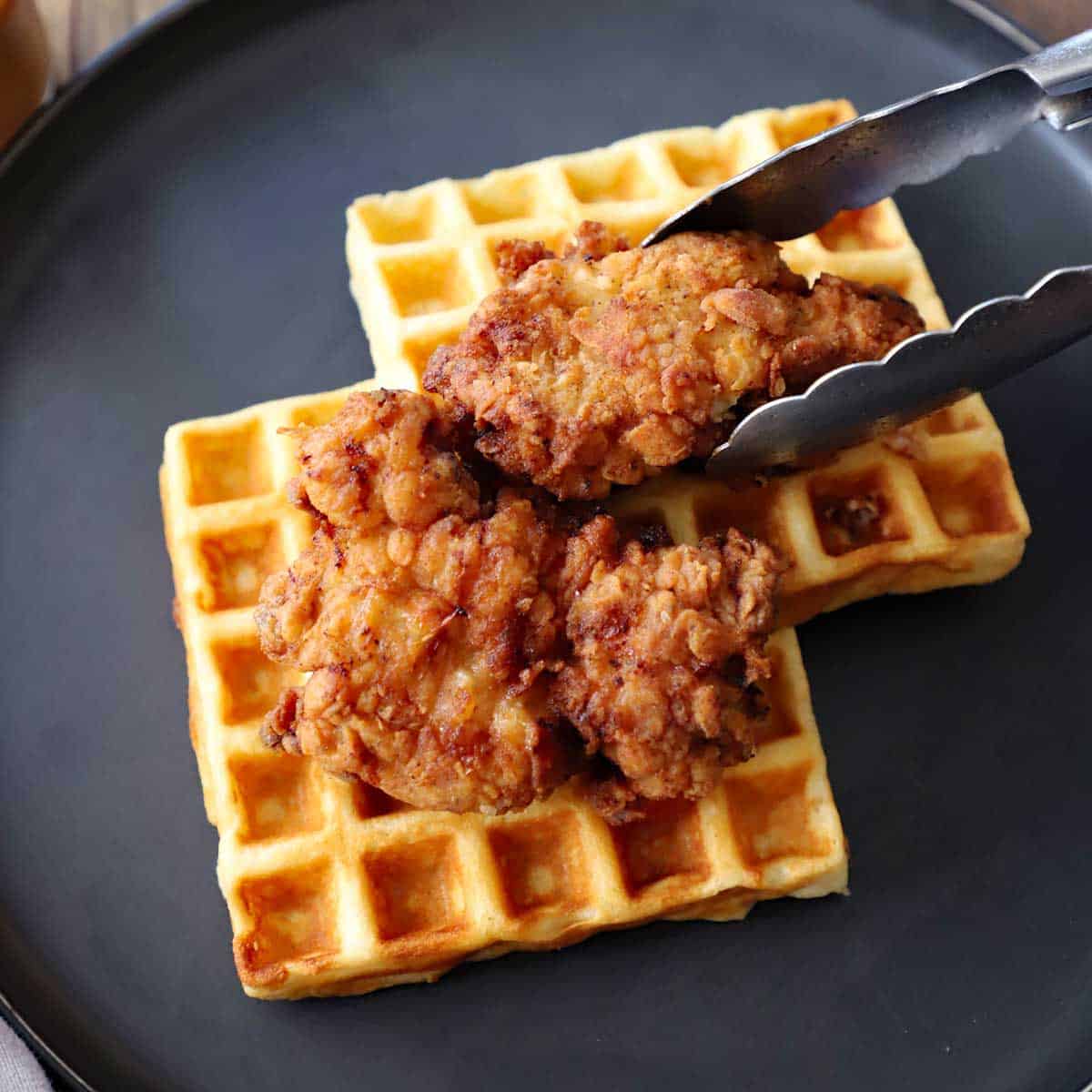 A person using a pair of metal tongs to place a crispy fried chicken piece on the top of two buttermilk waffles on a black dinner plate.
