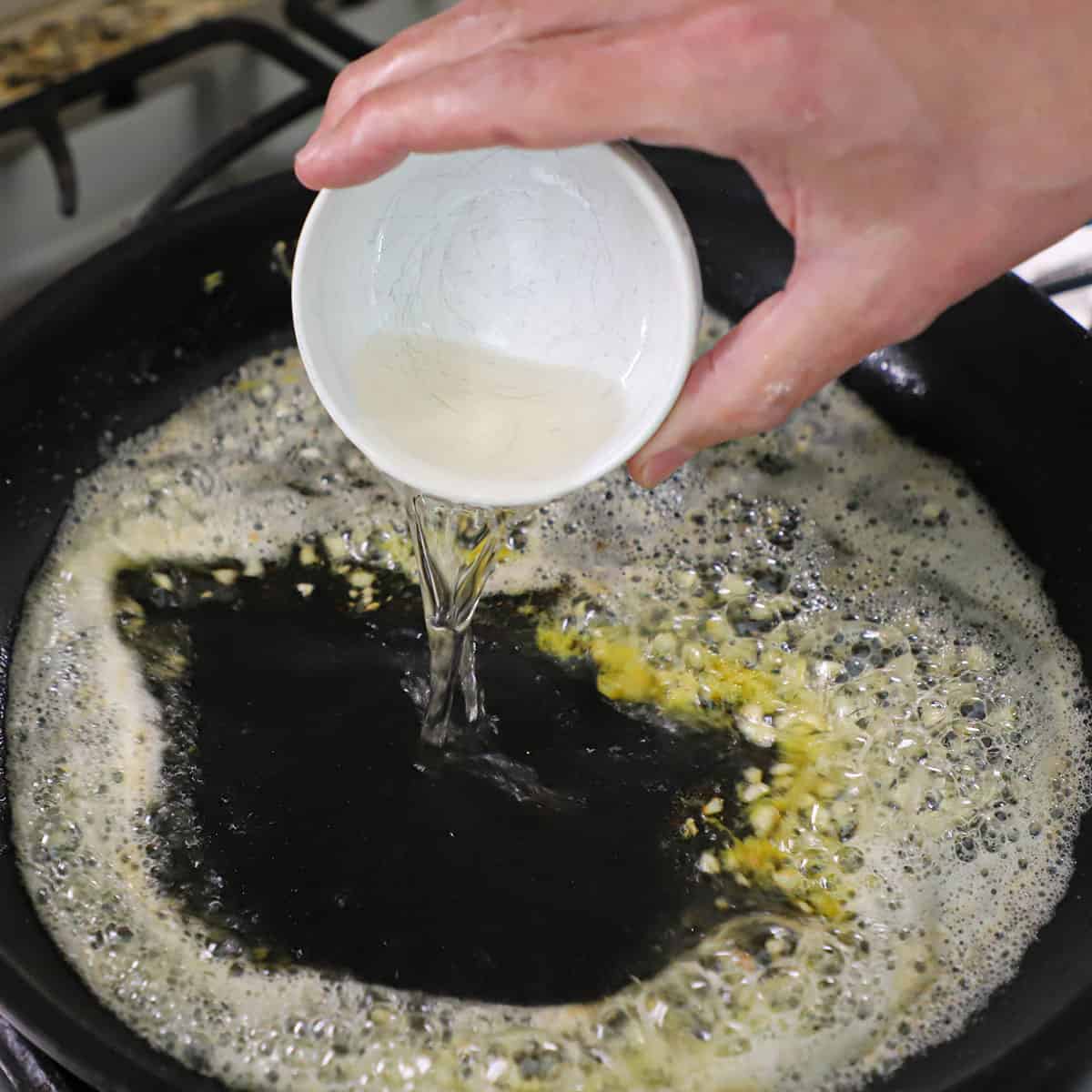 A person pouring white wine from a small white bowl into a black skillet filled with a simmering white wine, butter, and garlic sauce.