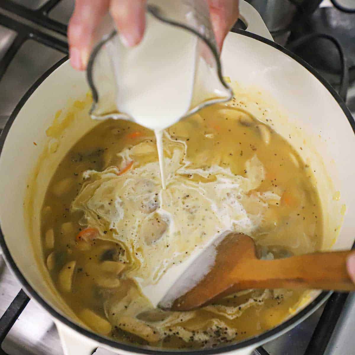 A person pouring heavy cream from glass measuring cup into a white pot that is filled with a brown gravy with sautéd mushrooms and vegetables.