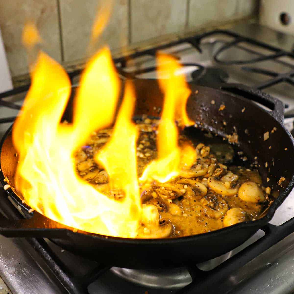 A large cast-iron skillet on a gas stove with a large flame filling much of the skillet as the mushrooms in the skillet are being flambéed.