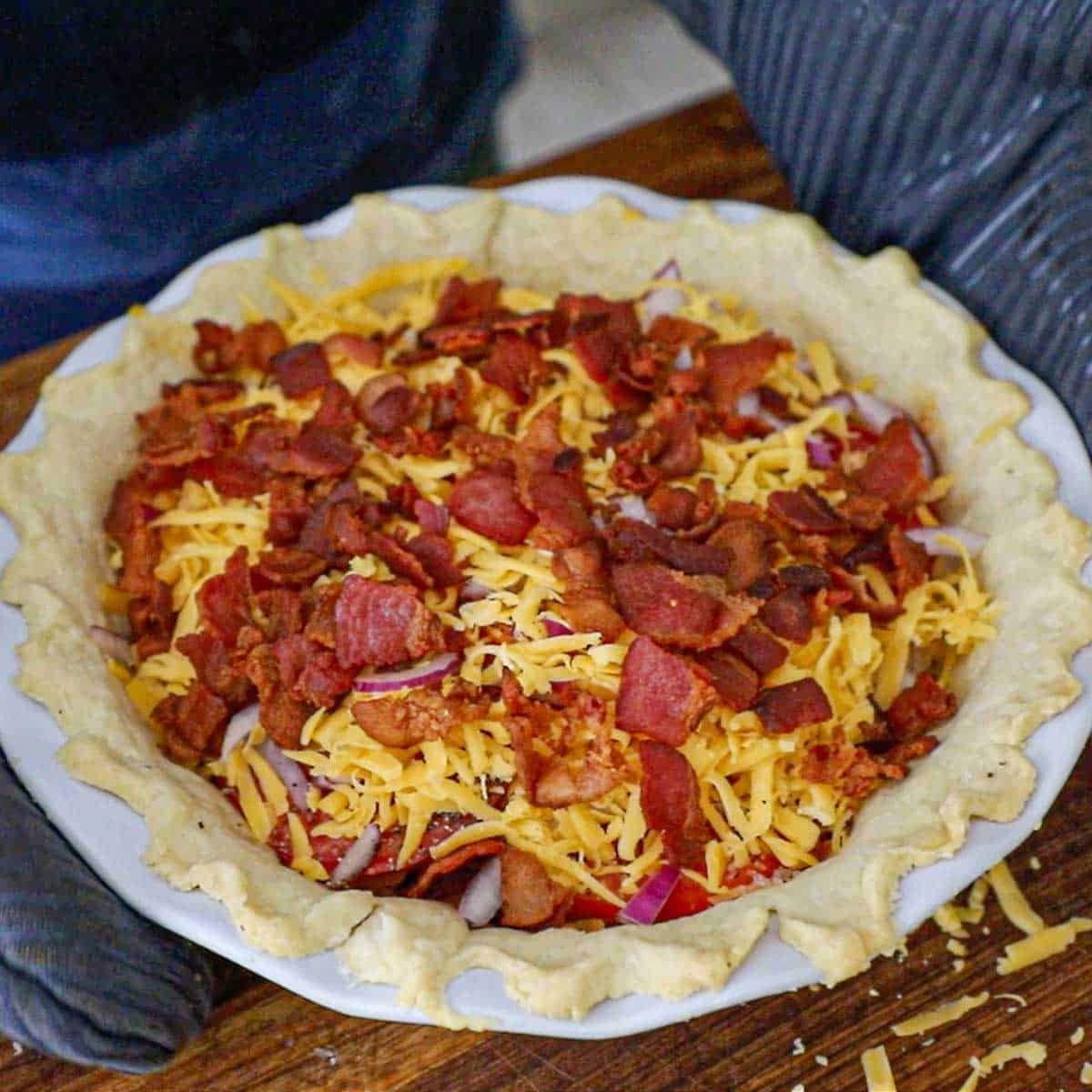 A person wearing oven mitts picking up a tomato cheddar and bacon pie that is in a white pie dish from a wooden cutting board.
