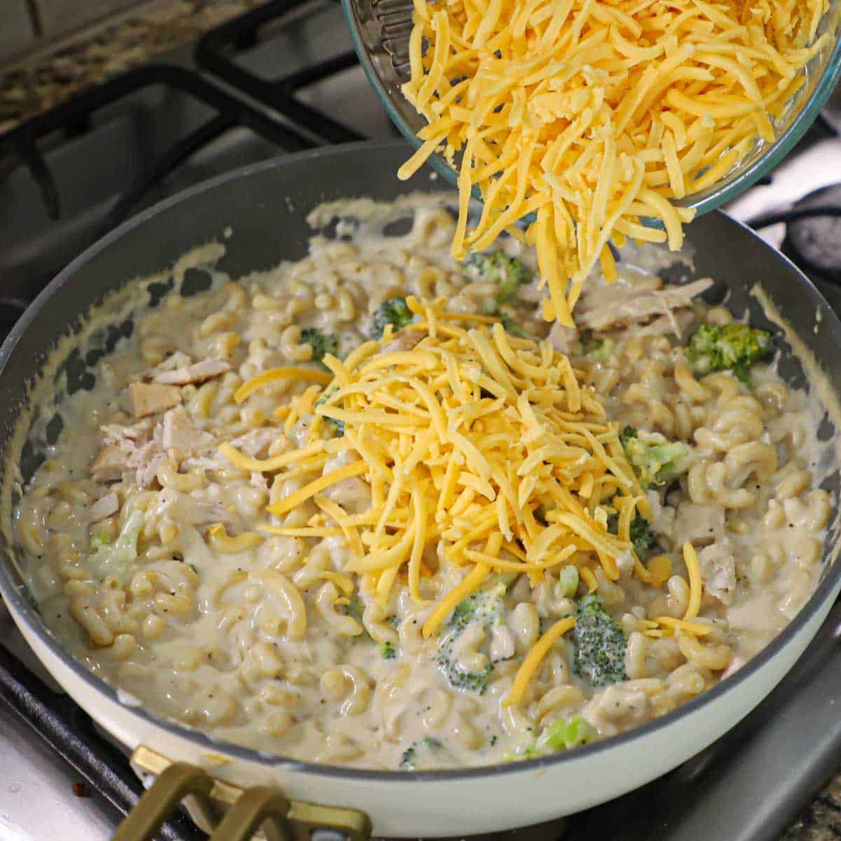 A person dumping shredded cheddar cheese and yellow American cheese from a glass bowl into a large skillet filled with creamy mac and cheese with chicken and broccoli.