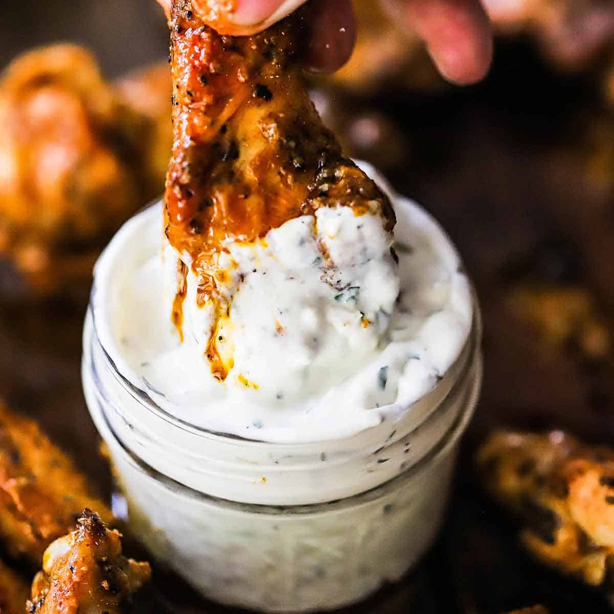 A person using his fingers to plunge a grilled chicken wing with buffalo sauce into a small jar filled with blue cheese dressing.