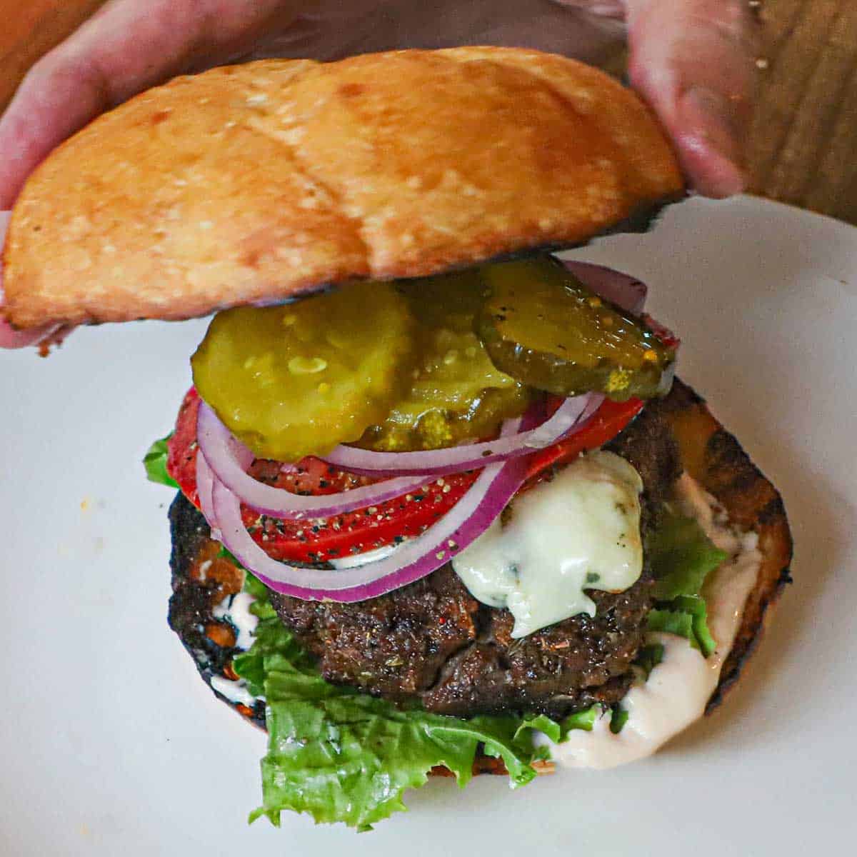 A person placing the top of a toasted hamburger bun on top of a black and bleu burger that consists of Cajun mayonnaise, lettuce, blackened burger, melty blue cheese, onions, lettuce, and pickles.