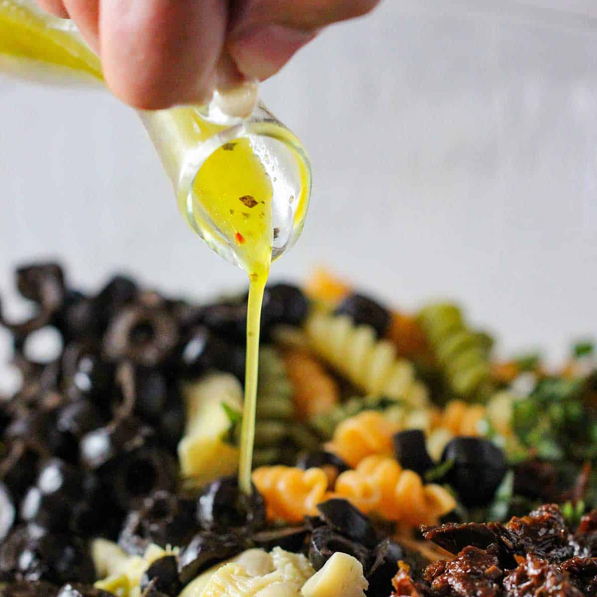 A person pouring homemade Italian dressing over the ingredients of a classic pasta salad in a bowl.
