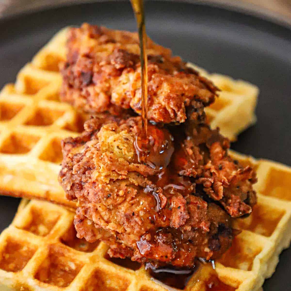 Maple syrup being poured over the top of a plate filled with crispy chicken and waffles.