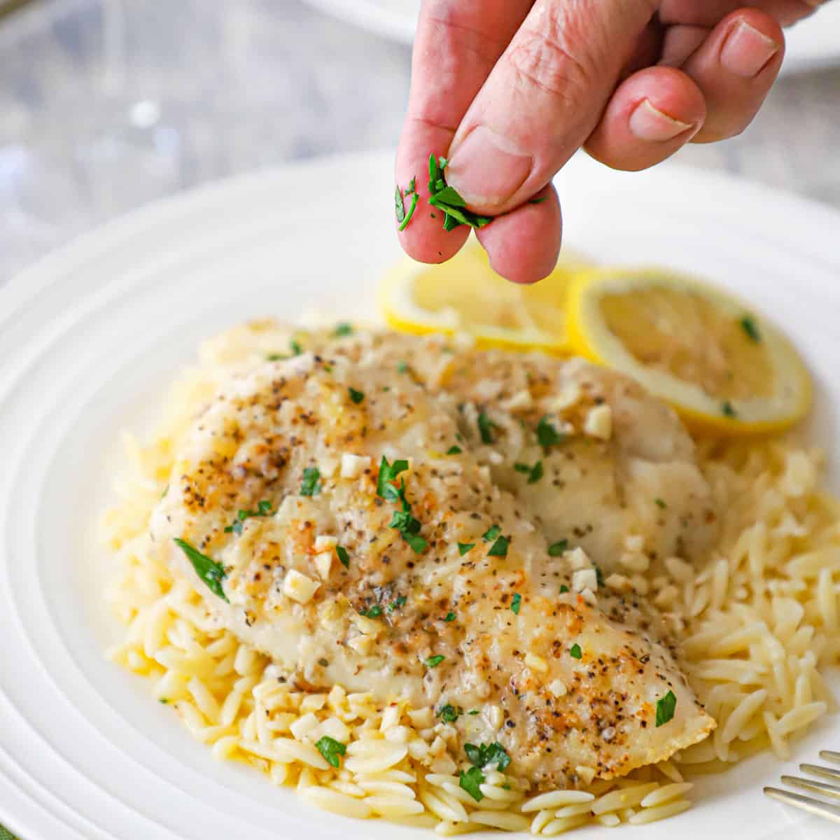A person using his fingers to sprinkle chopped Italian parsley over the top of a white dinner plate filled with a serving of pan-fried cod with lemon butter sauce.