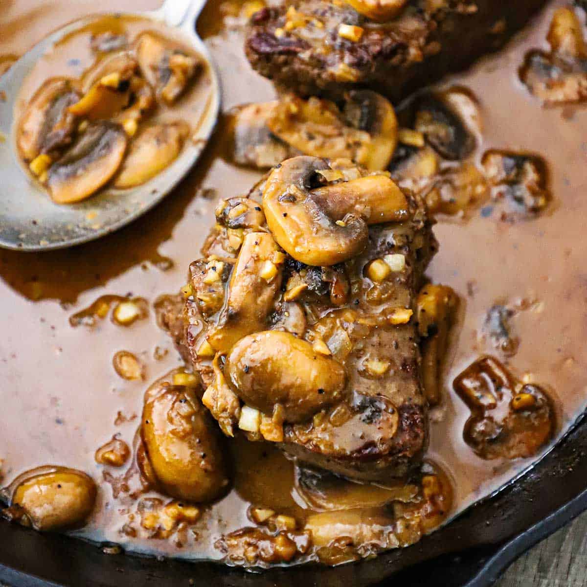 An overhead view of two filets of Classic Steak Diane in a cast-iron skillet that consists of seared filet mignon and sautéed mushrooms in a brown sauce.