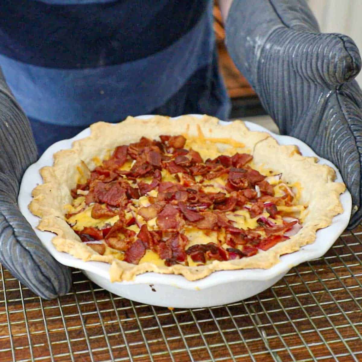 A person wearing oven mitts placing a freshly baked tomato cheddar and bacon pie onto a baking rack that is resting on a wooden cutting board.