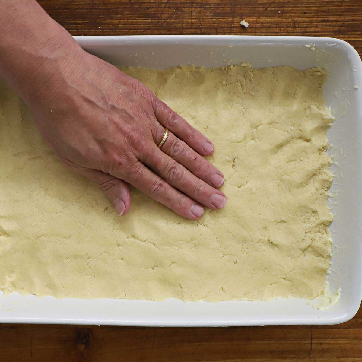 A person using his hand to press dough into the base of a white 9 by 13 inch baking dish.