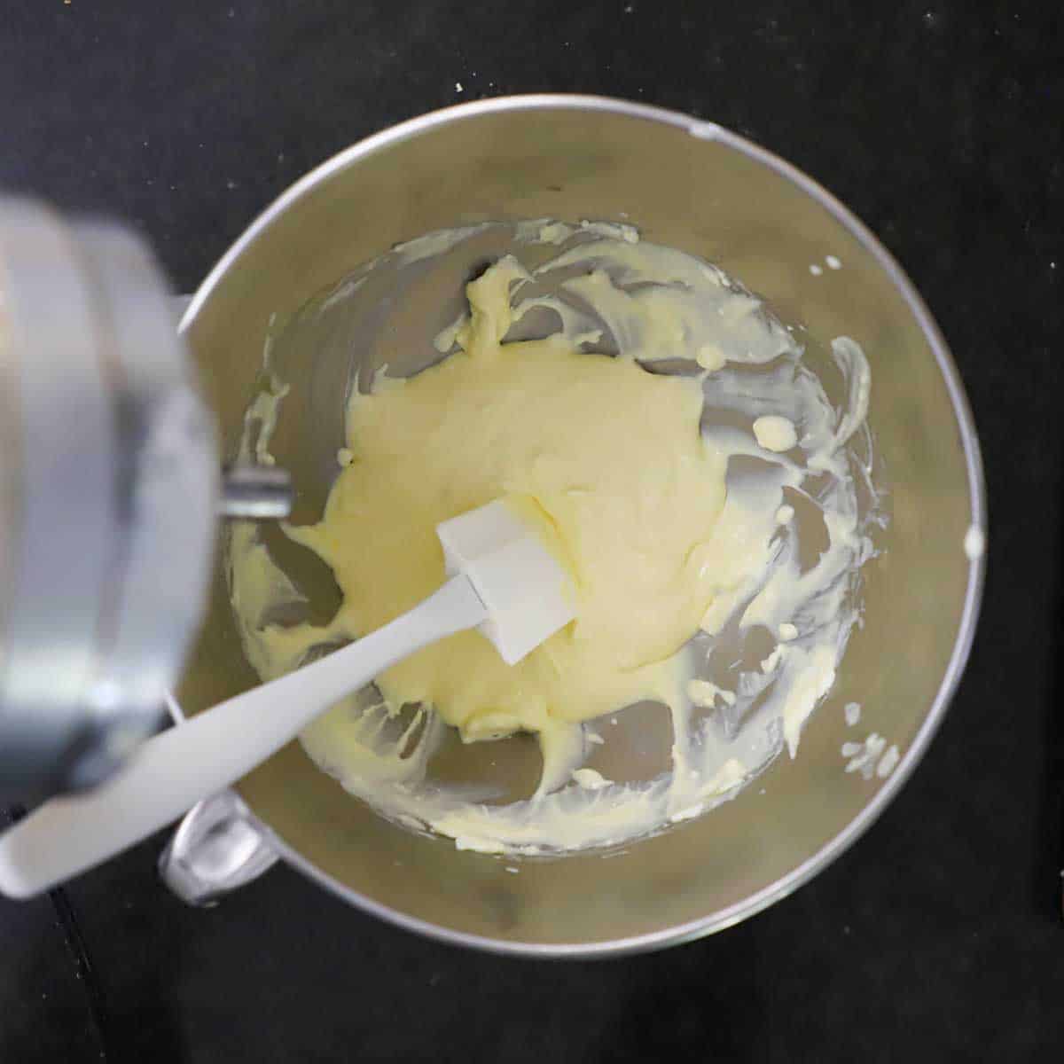 A white spatula resting in a stand-mixer bowl that is filled with softened cream cheese, eggs, and vanilla extract.