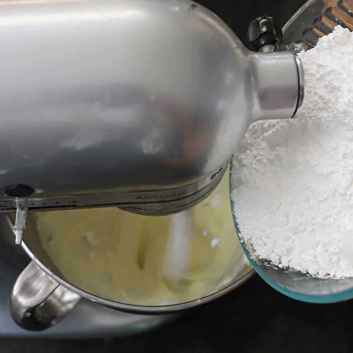 A person transferring powdered sugar from a glass bowl into a mixer that is beating a cream cheese mixture in the mixer bowl.