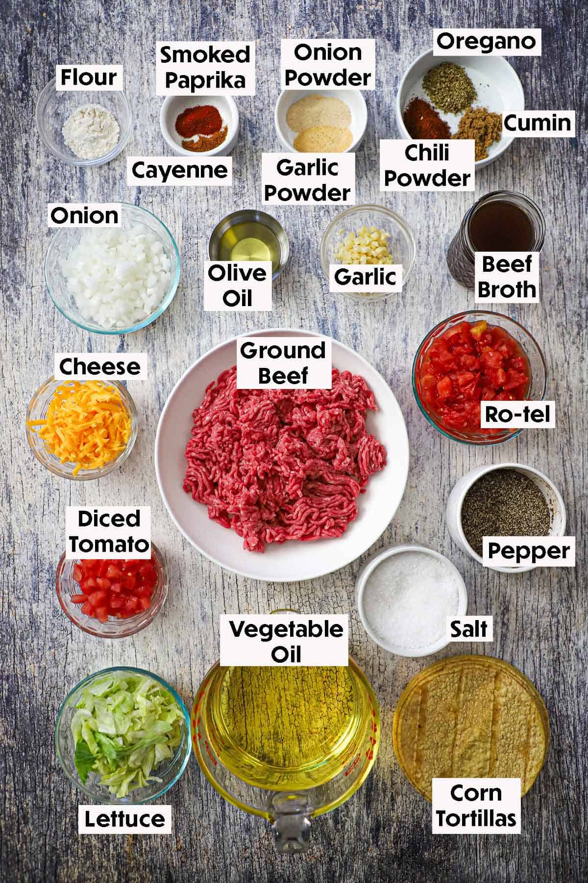 An arrangement of ingredients for Tex-Mex beef tacos on a grey wooden background including ground beef, onions,, garlic, beef broth, tomatoes, seasonings, cheese, lettuce, vegetable oil, and corn tortillas.