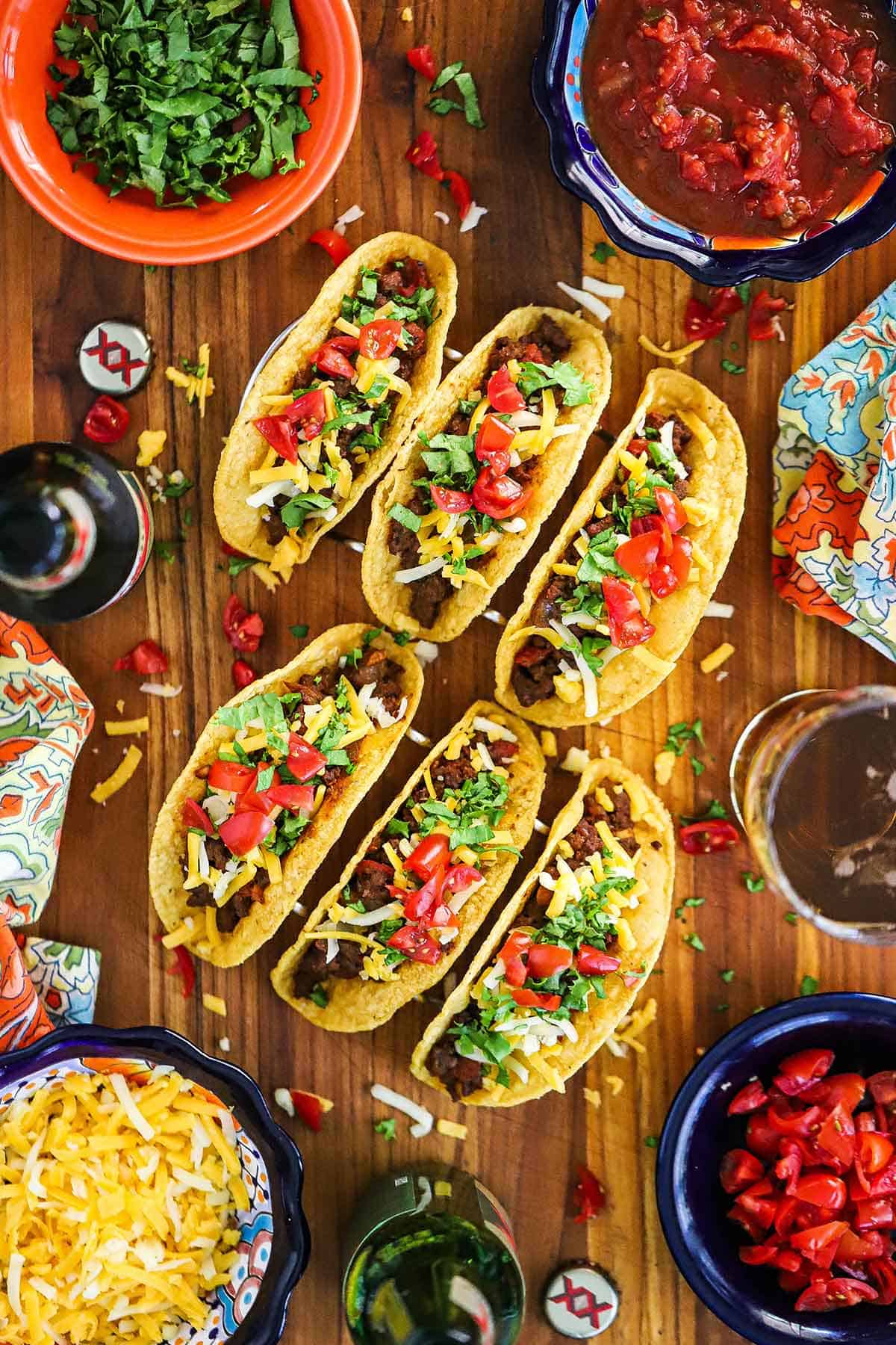 Two rows of crispy beef tacos resting on two taco holders with chopped tomatoes and shredded cheese scattered around them.