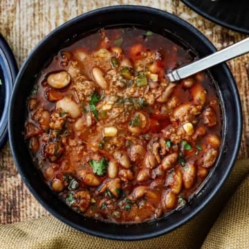 A dark blue soup bowl filled with a serving of chorizo and bean soup with a spoon resting in the middle of the soup bowl.