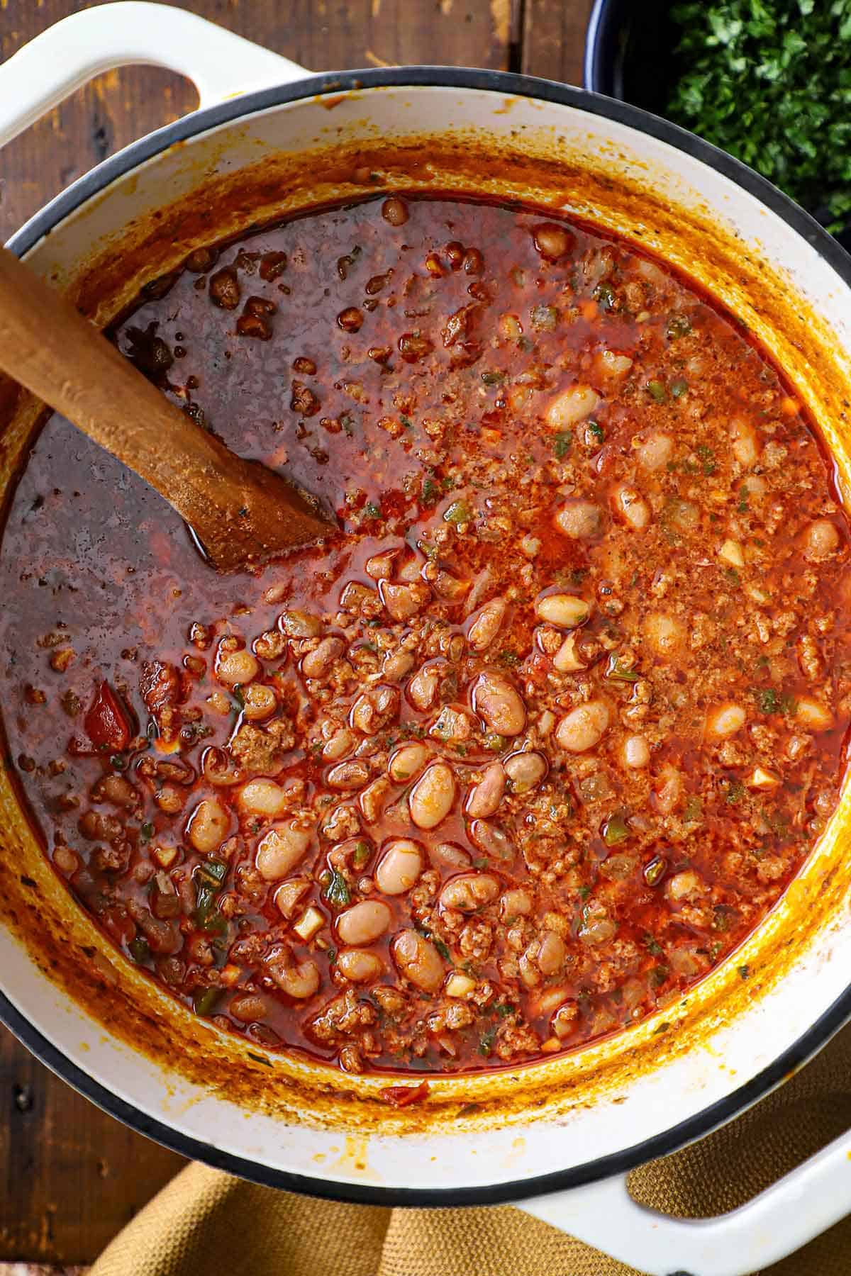 An overhead view of a large white pot filled with chorizo and bean soup with a wooden spoon in the middle of the soup.