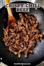 A black wok filled with freshly stir-fried crispy chili beef with a wooden spoon inserted off to the side.
