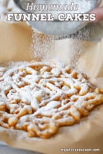 A person sifting powdered sugar from a sieve onto a freshly made homemade funnel cake that is resting on a brown piece paper.
