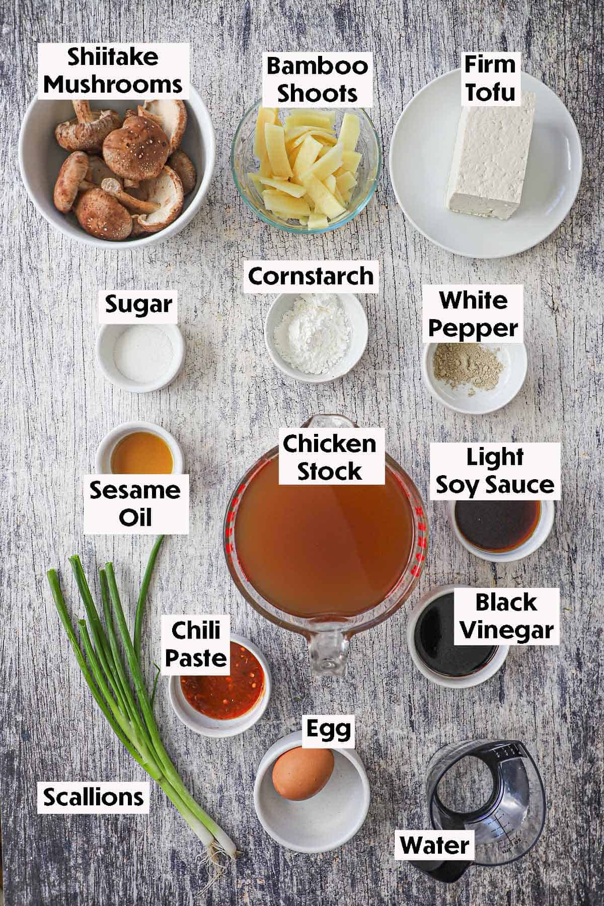 An arrangement of ingredients for homemade Chinese sweet and sour soup on wooden background including chicken stock, mushrooms, bamboo soots, tofu, seasonings, soy sauce, vinegar, chili paste, egg, and scallions.