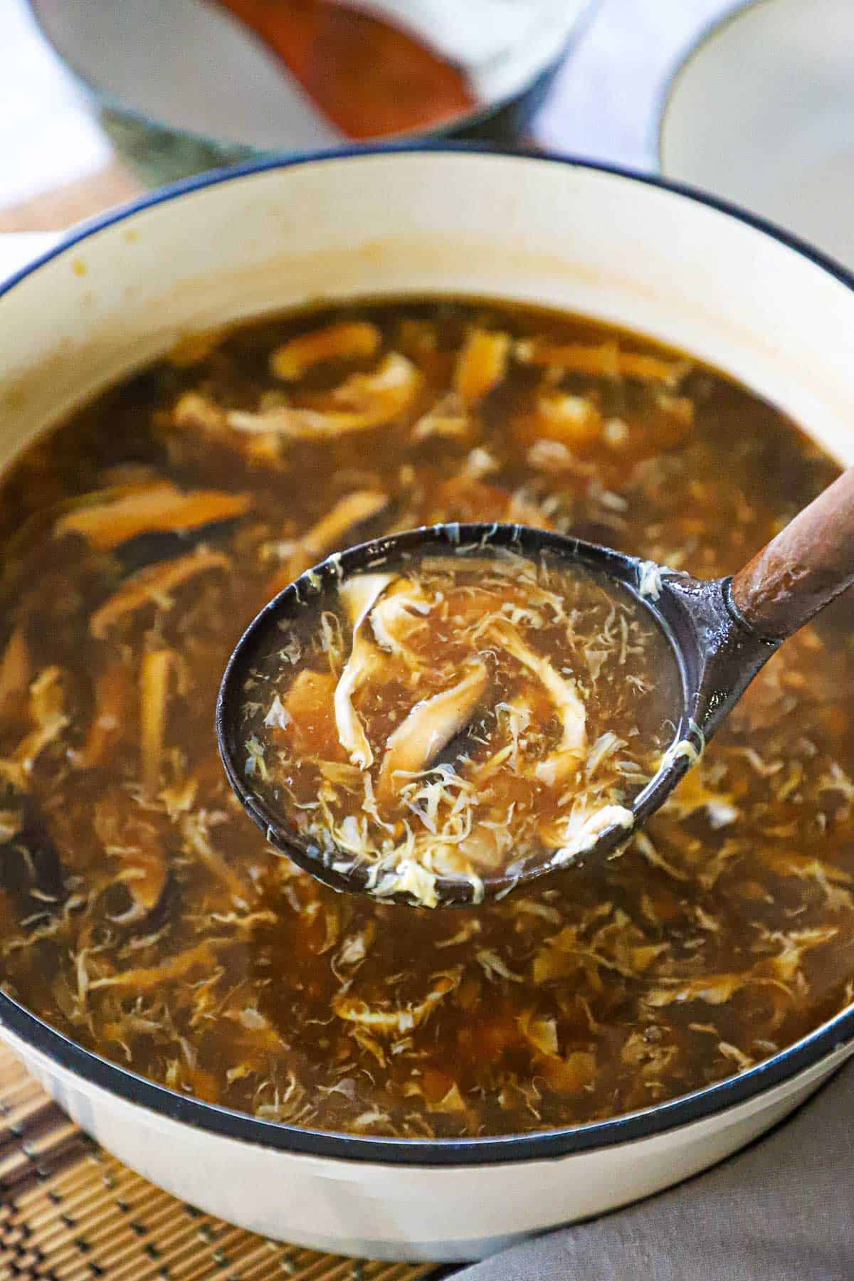 A large wooden ladle being used to raise a serving of homemade Chinese sweet and sour soup from a large white pot filled with the soup.