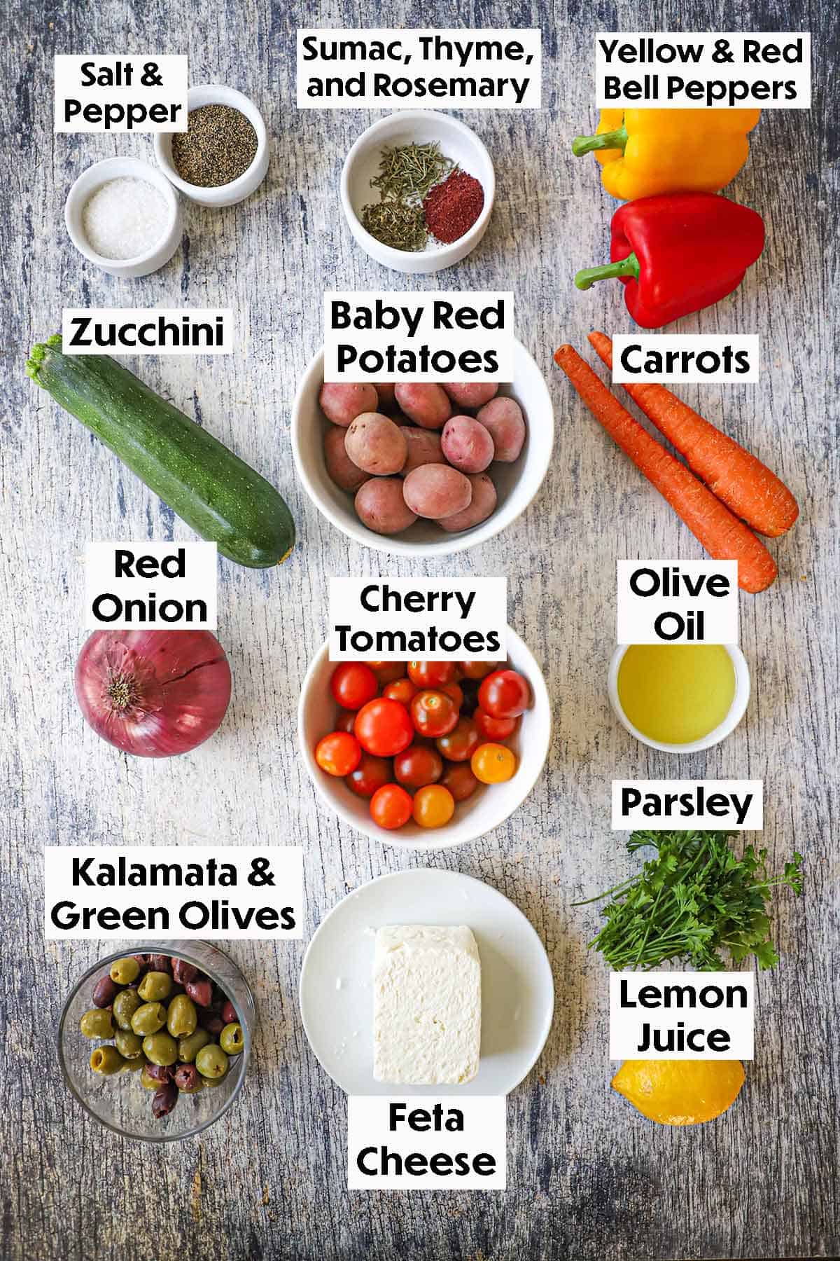 An arrangement of ingredients for Mediterranean roasted vegetables on a grey wooden background including potatoes, zucchini, bell peppers, tomatoes, onion, olives, herbs, olive oil, and feta cheese.