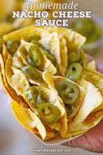 A person holding a paper basket filled with movie theater-style nachos topped with sliced jalapeño peppers.
