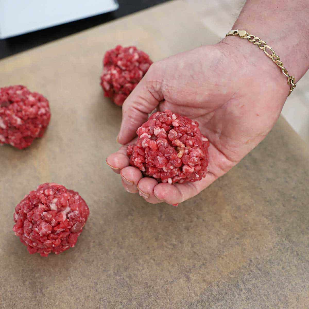 A person holding a round ball of ground beef in his hand over a piece of brown paper that is holding three other balls of beef.
