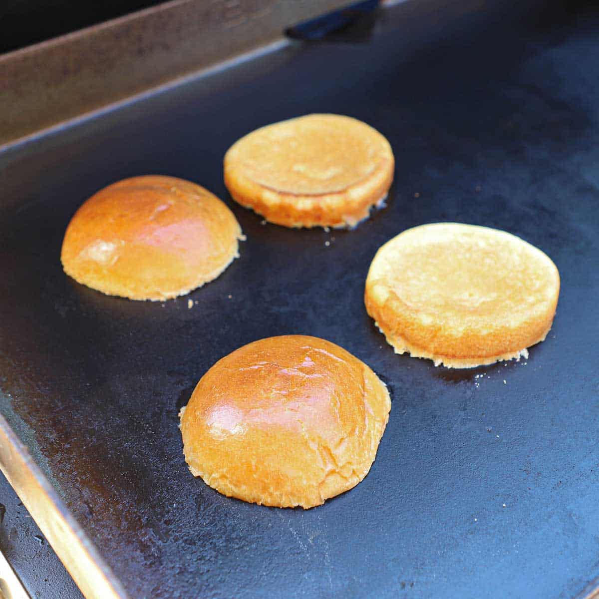 Four hamburger buns, two tops and two bottoms, being toasted cut-side down on a large outdoor griddle.