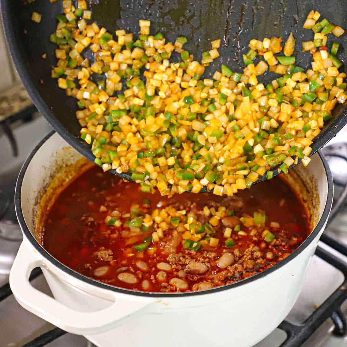 A person transferring sautéed chopped onions, jalapeños, and garlic from a skillet into a pot of simmering chorizo and bean soup.