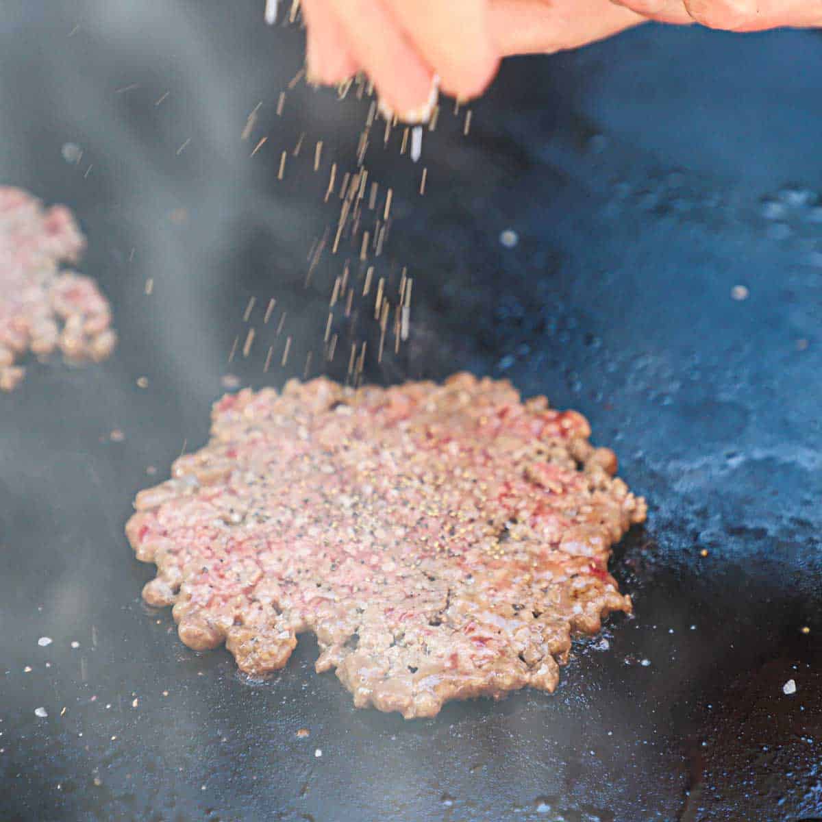 A person sprinkling salt and pepper onto a smash burger that is being cooked on an outdoor griddle.