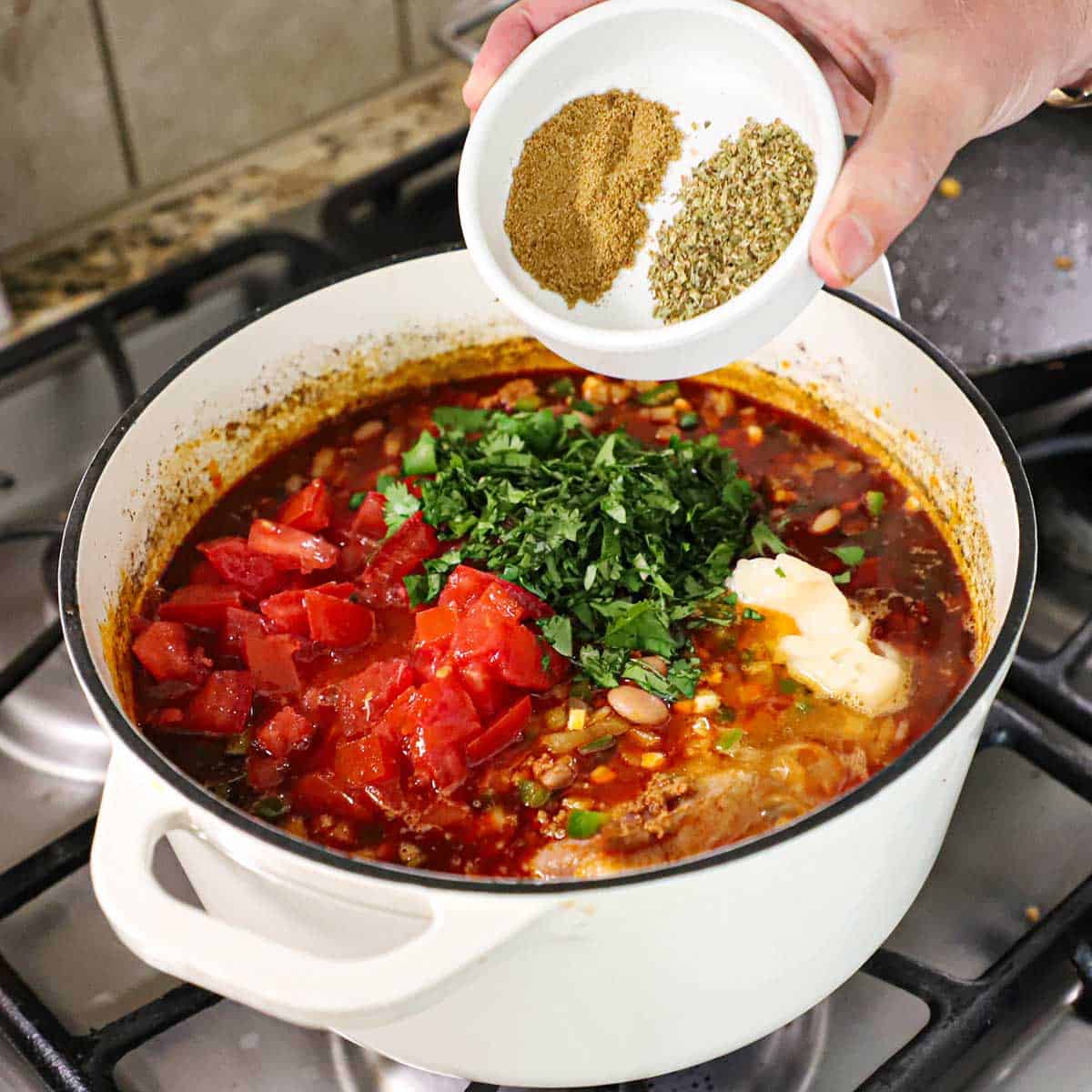 A person holding a small white bowl that is filled with a teaspoon each of dried cumin and dried oregano over a pot on a stove that is filled with a simmering chorizo and bean soup with chopped tomatoes and cilantro resting on the surface of the soup.