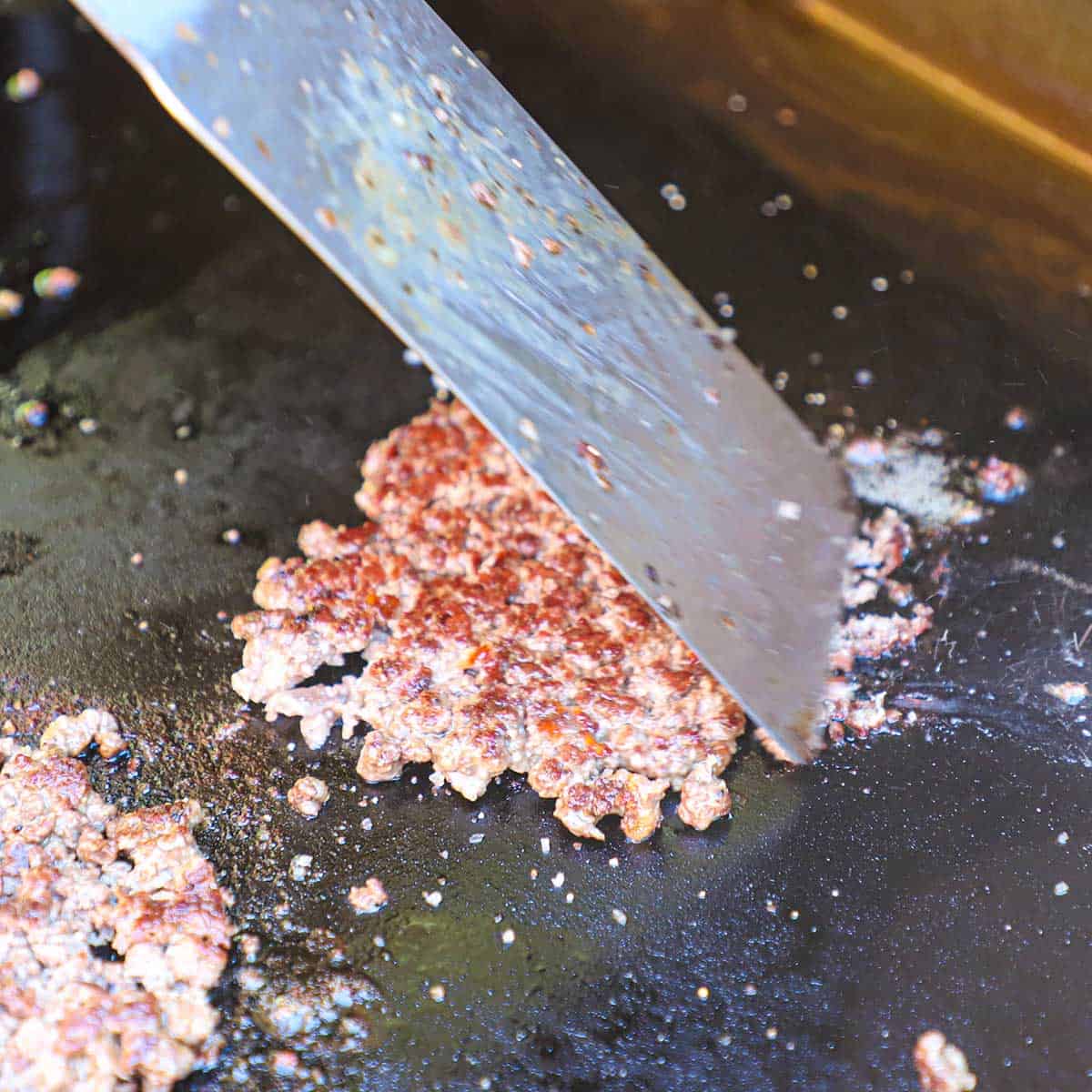 A person using a large spatula to push together a crispy smash burger patty that was just recently flipped on an outdoor griddle.