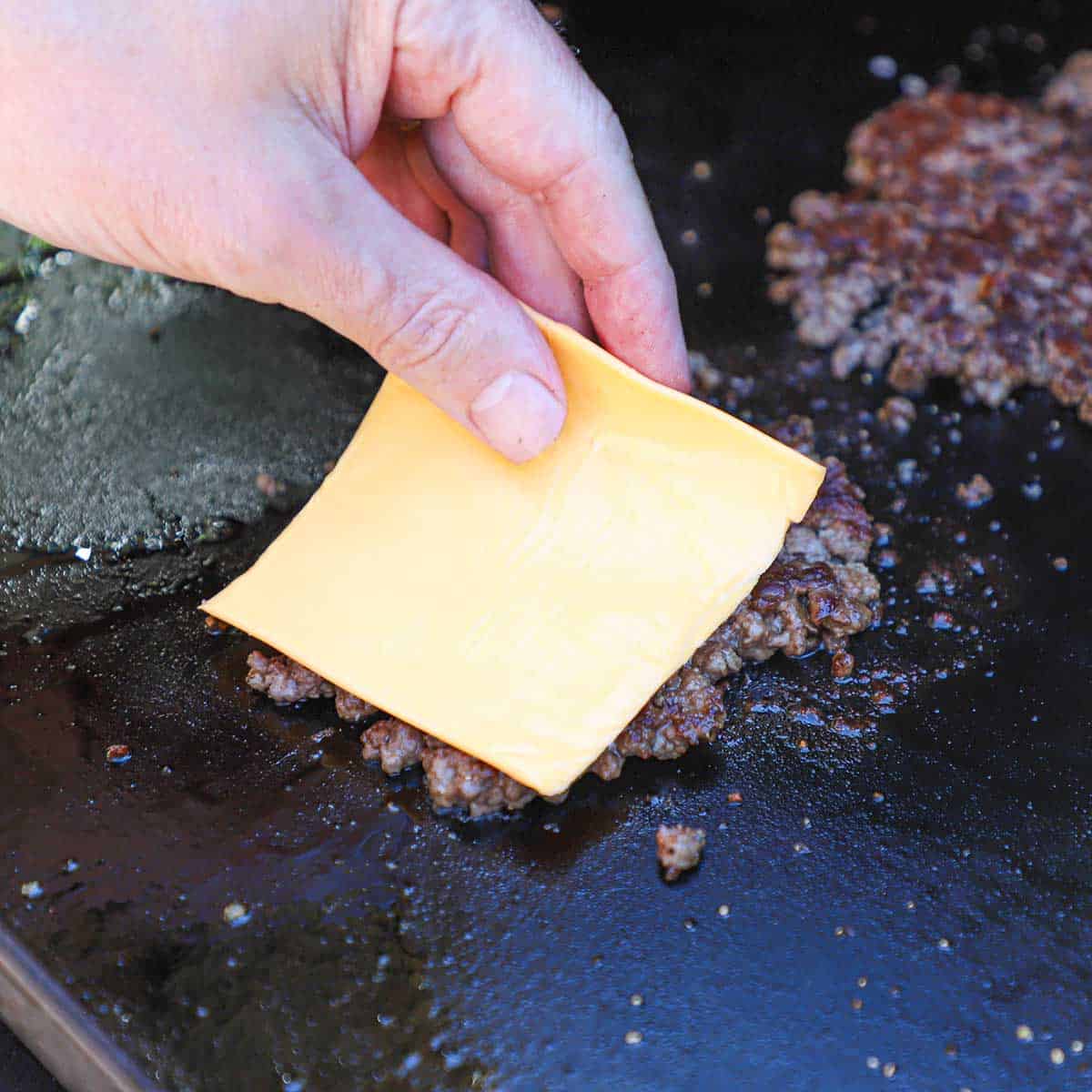 A person placing a square slice of yellow American cheese onto the top of a crispy smash burger that is sizzling on a large outdoor griddle.