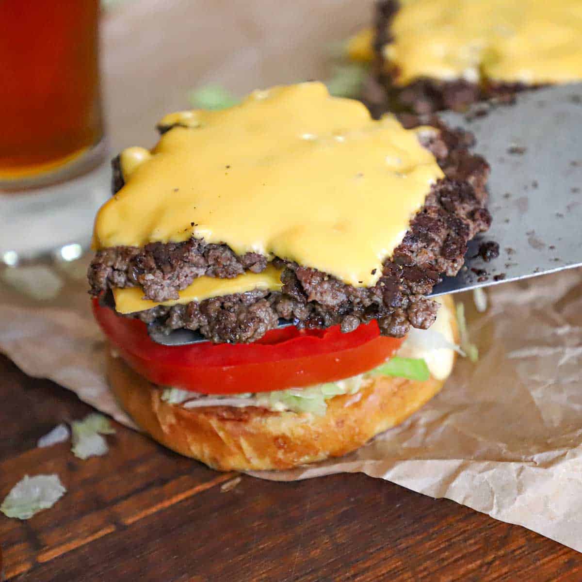 A person using a large metal spatula to place two smash burgers with American cheese onto a hamburger bun with burger sauce, lettuce, and tomato on it.