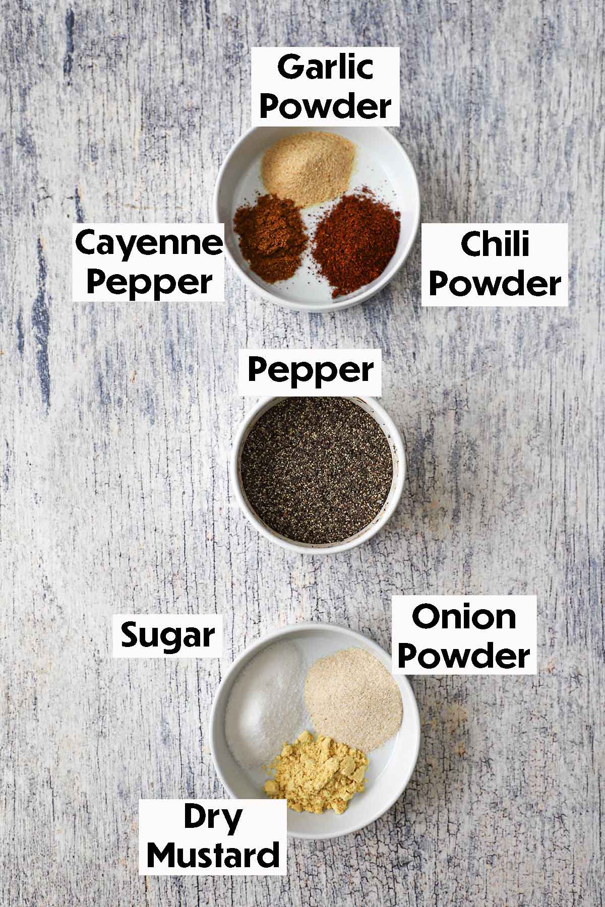 An arrangement of ingredients for Best Beef Rub on a grey wooden background including pepper, garlic powder, cayenne pepper, chili powder, sugar, onion powder, and dry mustard.
