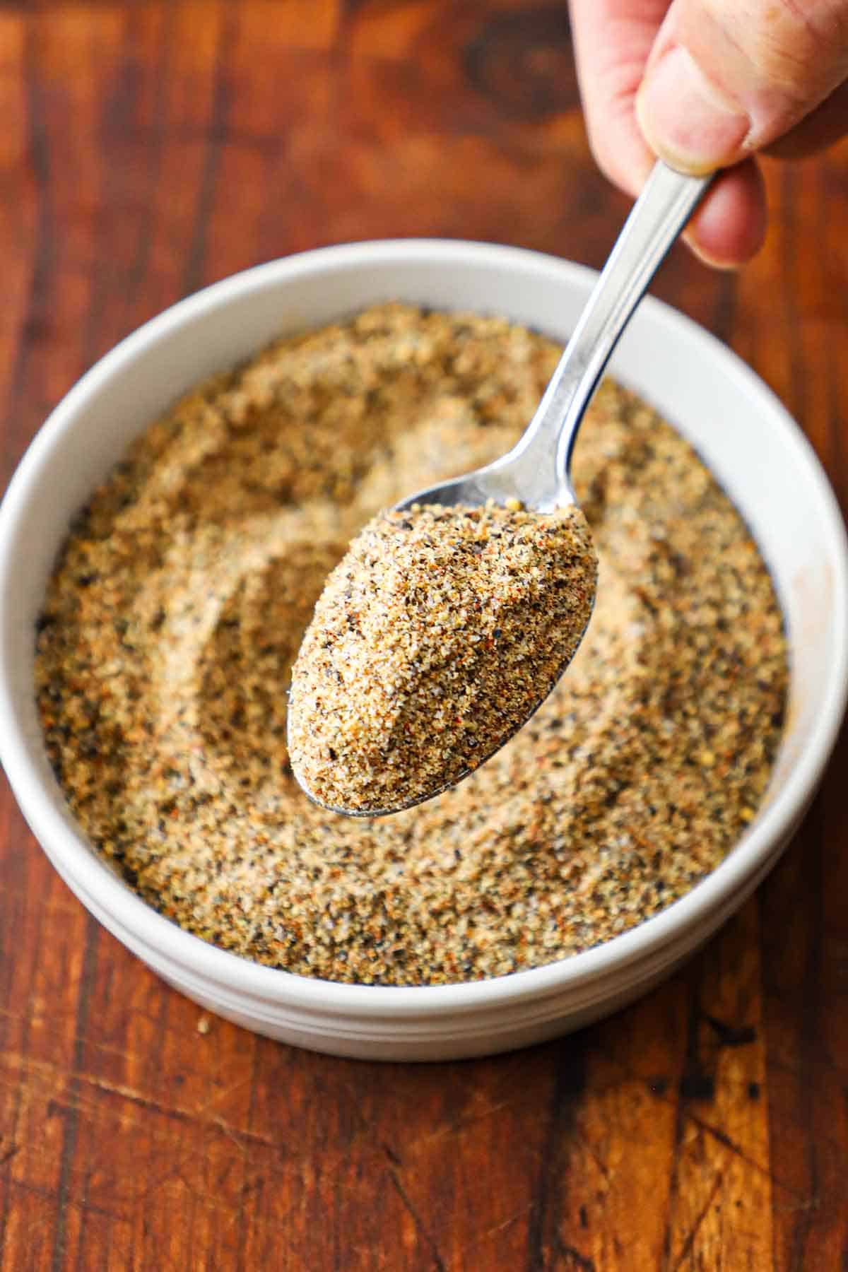 A person holding a spoon of Best Beef Rub over a small bowl filled with the same while resting on a wooden cutting board.