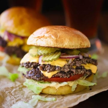 A close-up view of a double-stacked smash burger with melty cheese on each patty and garnished with pickles, red onion, shredded lettuce, and burger sauce.
